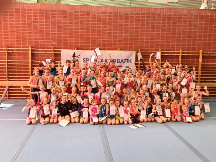Gruppenbild Große Gruppe junger Sportakrobat*innen posiert mit Urkunden und Medaillen in der Turnhalle.