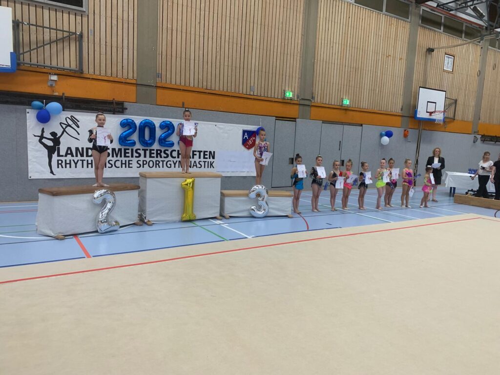 Siegerehrung bei einem Turnwettbewerb mit Podestplätzen und Teilnehmerinnen, die Urkunden halten.