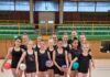Eine Gruppe von Gymnastinnen in schwarzer Sportkleidung, die bunte Bälle halten, steht in einer Turnhalle.