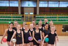 Eine Gruppe von Gymnastinnen in schwarzer Sportkleidung, die bunte Bälle halten, steht in einer Turnhalle.
