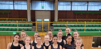 Eine Gruppe von Gymnastinnen in schwarzer Sportkleidung, die bunte Bälle halten, steht in einer Turnhalle.