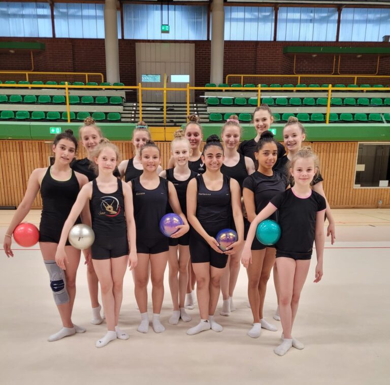Eine Gruppe von Gymnastinnen in schwarzer Sportkleidung, die bunte Bälle halten, steht in einer Turnhalle.