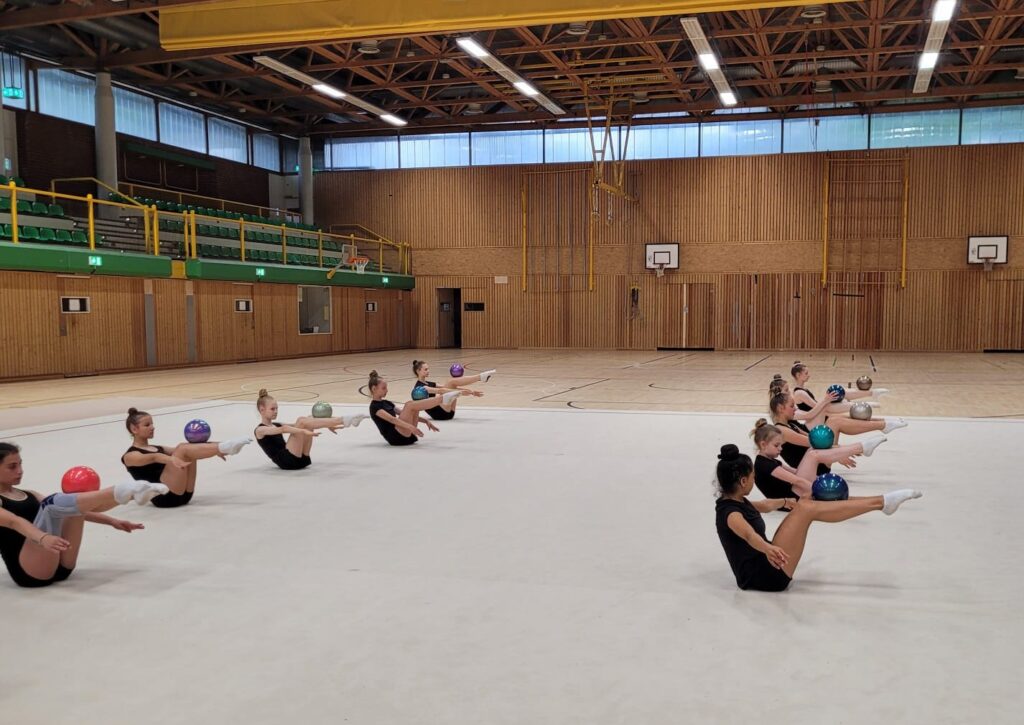 Eine Gruppe von Menschen in schwarzer Sportkleidung macht Gymnastikübungen in einer Turnhalle.